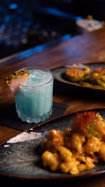 A pale blue cocktail beside shared plates at the bar